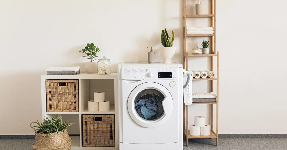 Mașină de spălat rufe frontală albă într-un spațiu de laundry luminos, cu plante și prosoape